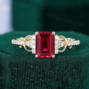 May include: A gold ring featuring a large, rectangular, red gemstone. The ring is embellished with smaller, clear gemstones and detailed goldwork on the band. The ring is displayed on a dark green velvet surface.