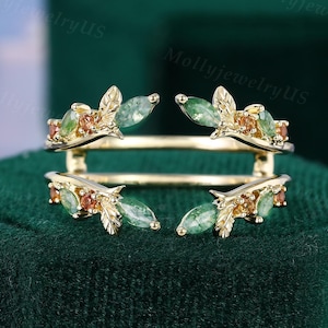 Puede incluir: Un anillo dorado con un diseño único. El anillo presenta piedras preciosas verdes y naranjas en forma de hoja. Está colocado sobre un fondo de terciopelo verde oscuro, con el texto "MollyjewelryUS" visible en el fondo.