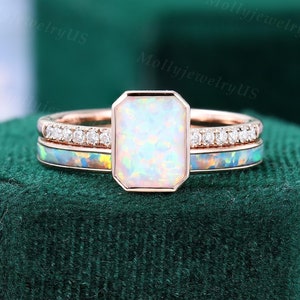 May include: A rose gold engagement ring set with a large, rectangular, white opal gemstone. The ring is surrounded by a band of smaller diamonds and two additional bands, one with a white opal inlay and the other with a blue opal inlay.