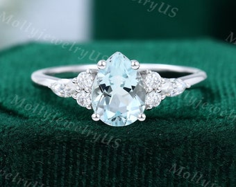 May include: A silver ring featuring a pear-shaped, light blue gemstone as the centerpiece, flanked by smaller, round, clear gemstones. The ring is set against a dark green velvet background, highlighting the ring's design and color.