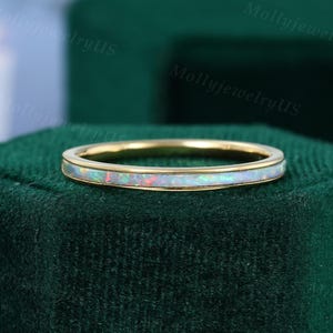 Può includere: Un anello a fascia in oro con un profilo sottile. L'anello presenta un intarsio di opale bianco con lampi di colore. L'anello è posto su uno sfondo di velluto verde scuro.