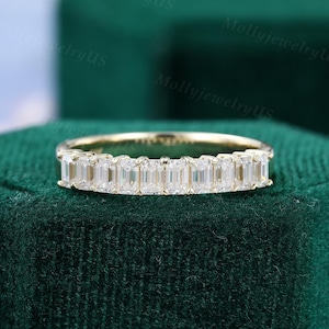 Puede incluir: Un anillo de banda de oro con una fila de diamantes de talla esmeralda. El anillo está sobre una superficie de terciopelo verde.