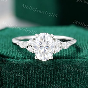 May include: A silver-toned ring featuring a large oval-cut center stone flanked by two smaller round stones and one pear-shaped stone on each side. The band is thin and polished, showcasing a multi-stone arrangement that creates a symmetrical design.