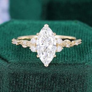 May include: A gold ring featuring a large marquise-cut center stone flanked by three small round stones on each side. The band has a detailed vine-like texture with small embedded diamonds extending along the sides of the metal shank.