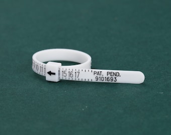May include: A white plastic ring sizer with black markings and text. The ring sizer has a band with numbers and an arrow, and a measuring strip with the text "PAT. PEND. 9101693". The ring sizer is on a dark green surface.