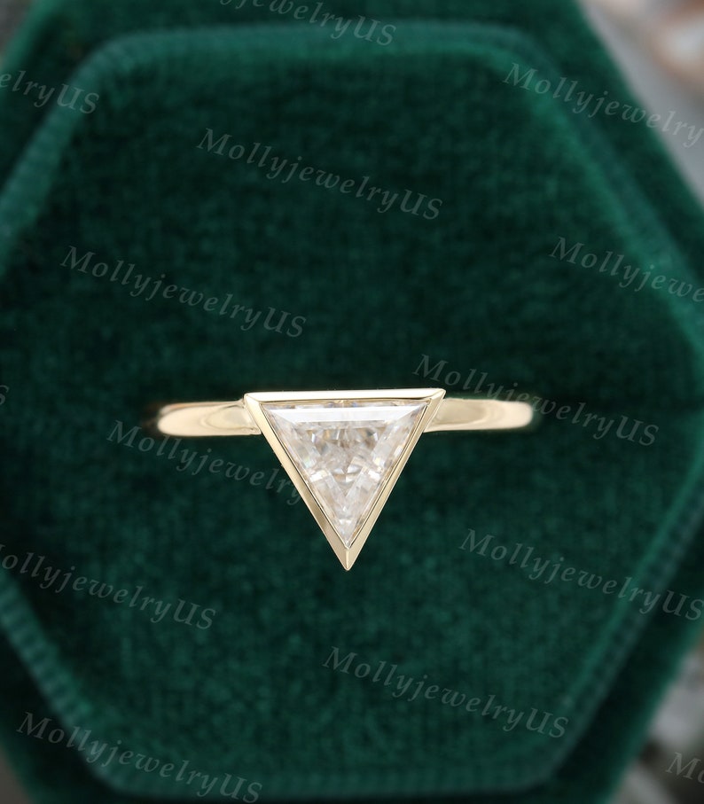 Pu&ograve; includere: Un anello di fidanzamento in oro con un diamante di forma triangolare incastonato a griffe. L'anello &egrave; esposto in una scatola per anelli in velluto verde.