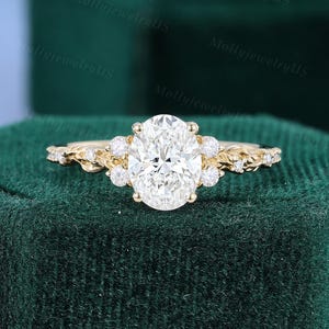 Peut inclure: Une bague de fiançailles en or avec une pierre centrale en diamant de taille ovale. La bague présente de plus petits diamants et un motif floral sur le bracelet. La bague est posée sur un fond vert foncé.