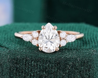 May include: Rose gold engagement ring featuring a pear-shaped diamond center stone, flanked by smaller round and pear-shaped diamonds. The ring is set on a delicate band, showcasing a classic and elegant design. The ring is displayed on a dark green velvet surface.