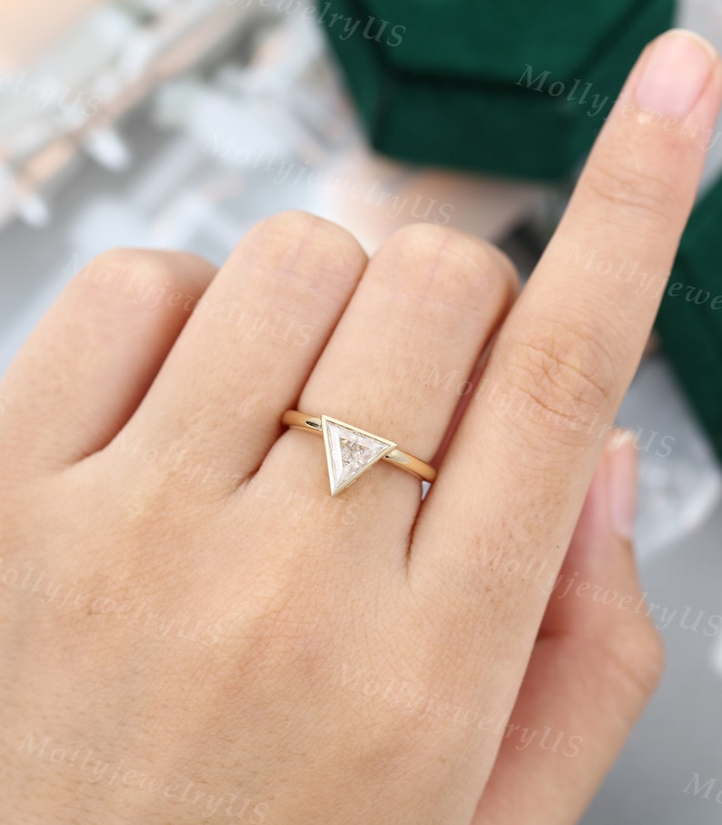 Pu&ograve; includere: Un anello d'oro con un diamante di forma triangolare incastonato in una montatura a bezel. L'anello &egrave; su un dito.