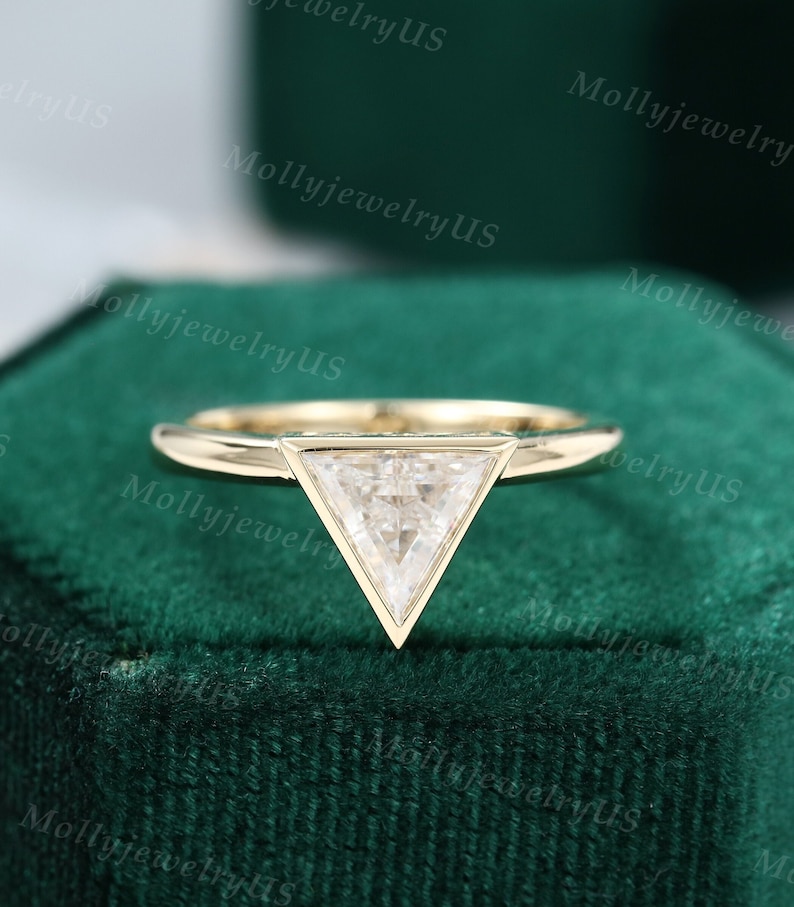 Pu&ograve; includere: Un anello di fidanzamento in oro con un diamante triangolare incastonato a griffe. L'anello &egrave; esposto in una scatola per anelli in velluto verde.