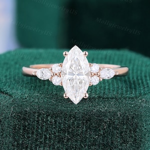 May include: A rose gold engagement ring featuring a marquise-cut diamond center stone, flanked by smaller round and pear-shaped diamonds. The ring is set on a dark green velvet surface.