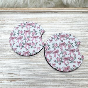 May include: Two car coasters with a floral and bow design. The coasters feature a white background with pink roses and bows. The edges are black, and the coasters have a semi-circle cut-out.