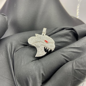 May include: A silver pendant in the shape of a panther's head, encrusted with small, clear stones. The panther has red eyes and a detailed, open mouth. The pendant is suspended from a small loop at the top.