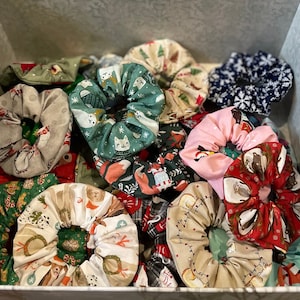 May include: A collection of fabric hair scrunchies featuring various holiday-themed patterns including gingerbread men, snowflakes, cats, foxes, and festive lights in colors such as red, green, blue, pink, and beige, all gathered together in a box.