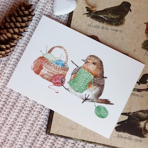 May include: A watercolor painting of a robin bird knitting with yarn and needles. The bird is sitting next to a basket of yarn and needles. The painting is on a white background.