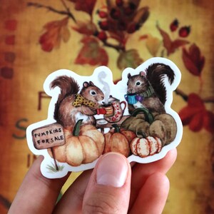 May include: Two squirrels wearing scarves and holding mugs sit on a table with pumpkins and a sign that reads "Pumpkins for Sale".