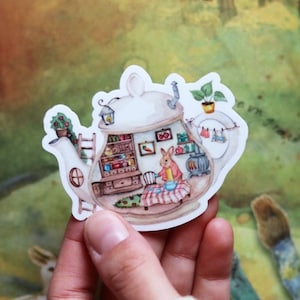 Può includere: Un adesivo stravagante raffigurante una casa a forma di teiera con un coniglio all'interno. L'illustrazione include un interno accogliente con mobili, una stufa e un piccolo giardino. L'adesivo è tenuto su uno sfondo sfocato con toni verdi e marroni.