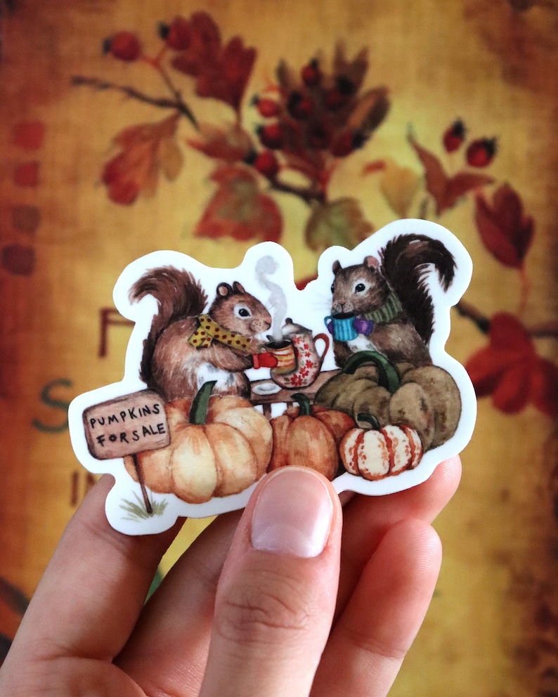 May include: Two squirrels wearing scarves and holding mugs sit on a table with pumpkins and a sign that reads "Pumpkins for Sale".