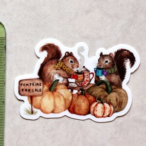 May include: Two brown squirrels wearing scarves and holding mugs sit on a table with pumpkins and a sign that reads "Pumpkins for Sale".