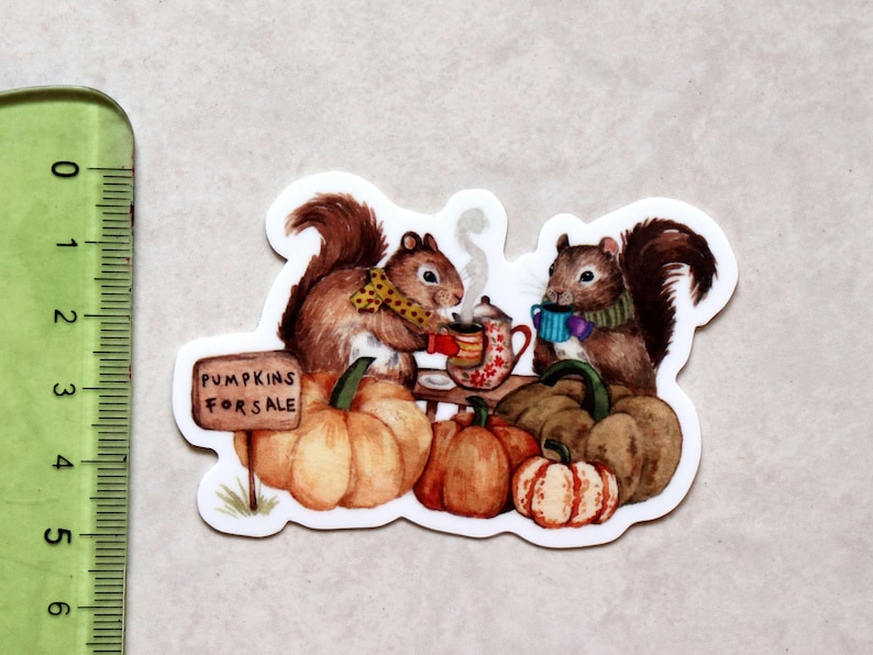May include: Two brown squirrels wearing scarves and holding mugs sit on a table with pumpkins and a sign that reads "Pumpkins for Sale".