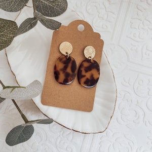 May include: A pair of gold and brown tortoise shell earrings. The earrings feature a small gold disc with a textured pattern and a larger oval-shaped tortoise shell pendant.