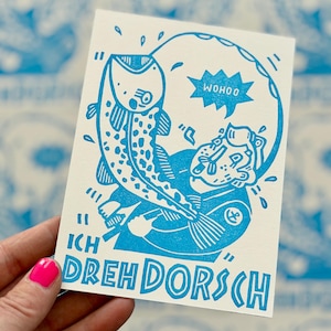 Könnte beinhalten: Eine rechteckige weiße Karte mit einer blauen Tintenillustration einer Person, die einen großen Fisch hält. Der Text WOHOO erscheint in einer Sprechblase und der Text ICH DREH DORSCH ist unten aufgedruckt. Das Design verwendet kräftige Linien.