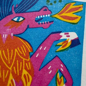 以下が含まれることがあります： 火を吹くドラゴンのカラフルなイラスト。 ドラゴンは、青い背景にピンク、オレンジ、黄色の色合いで描かれています。 このアートワークは、大胆な線とテクスチャのある外観を持つリノカットスタイルです。