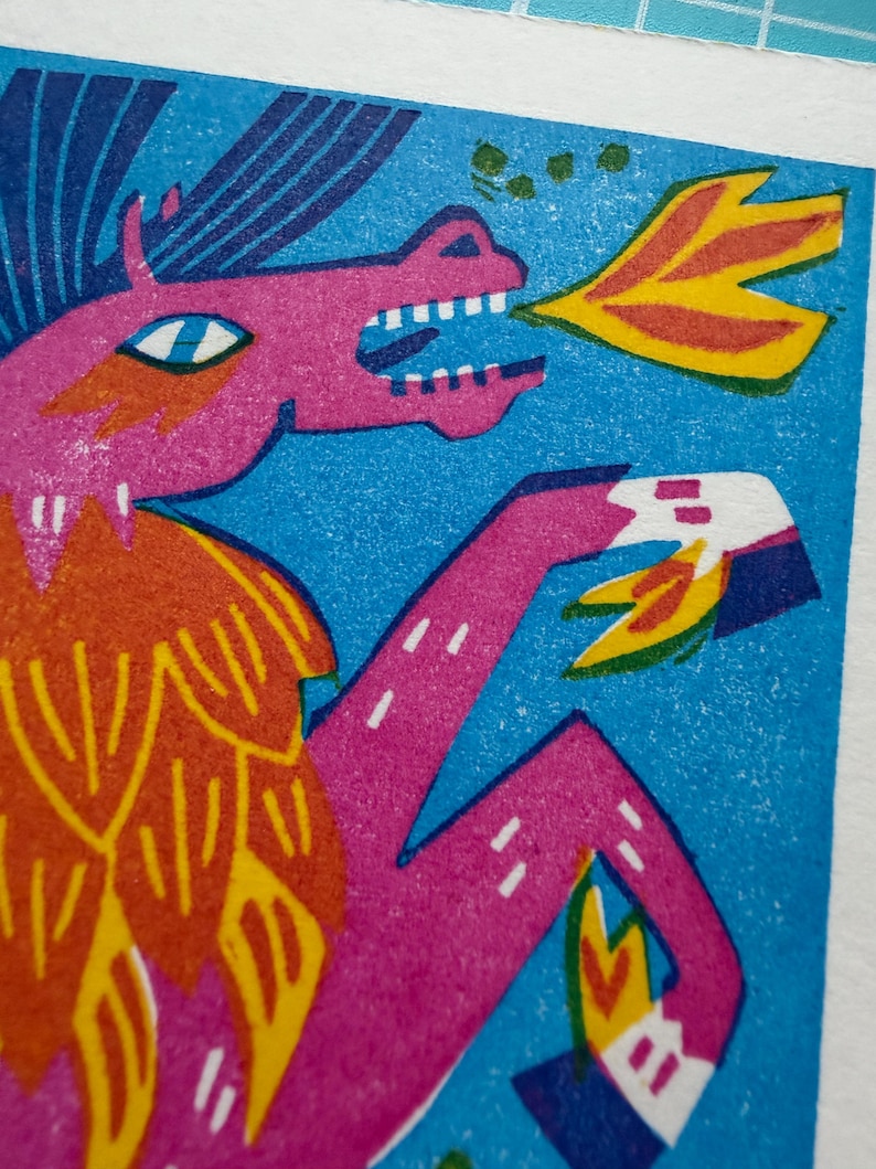 以下が含まれることがあります： 火を吹くドラゴンのカラフルなイラスト。 ドラゴンは、青い背景にピンク、オレンジ、黄色の色合いで描かれています。 このアートワークは、大胆な線とテクスチャのある外観を持つリノカットスタイルです。