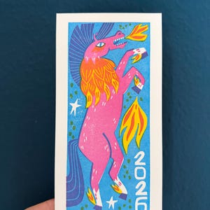 以下が含まれることがあります： 炎のたてがみと尾を持つピンク色のユニコーンのイラスト。背景は青色で、星と「2026」の文字が描かれています。ユニコーンは立ち上がり、口から炎が出ています。長方形の白いカードに描かれたアートワークです。