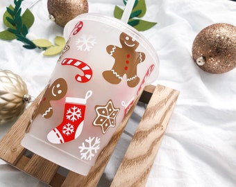 May include: White reusable cup with a frosted finish featuring a Christmas-themed design. The design includes gingerbread men, candy canes, snowflakes, and stockings in red, white, and brown.