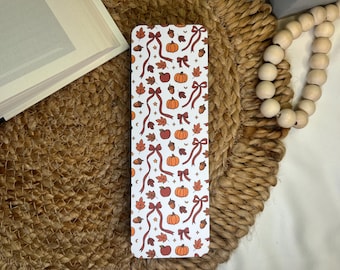 May include: A rectangular bookmark with a white background, adorned with a repeating pattern of orange pumpkins, red apples, brown acorns, and autumn leaves. Long, flowing ribbons in a deep red color add a decorative touch. The bookmark is a fall-themed item.