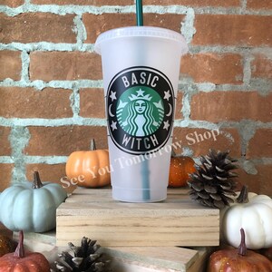 May include: A clear plastic reusable cup with a black and green design that says "Basic Witch" with a mermaid-like figure and stars. The cup is sitting on a wooden surface with autumnal decorations.