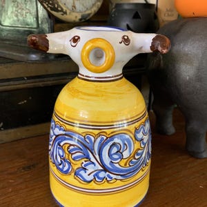 May include: A yellow ceramic pitcher with a whimsical design. The pitcher has a round body with a blue and white floral pattern. The spout is shaped like a ring and the handle is shaped like a cow's head.