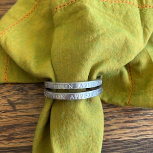 May include: A green linen napkin with orange stitching is held by two silver napkin rings. The rings are engraved with the words "BUON APPETITO". The napkin is folded and draped, creating a textured appearance.