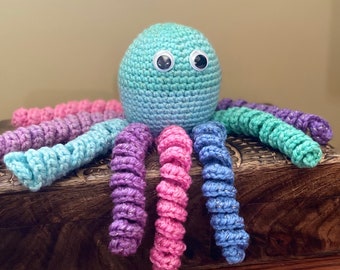May include: A crocheted octopus toy with a blue, green, and purple ombre body and eight curled tentacles. The tentacles are a mix of pink, purple, and blue with glitter accents. The octopus has two large black eyes.