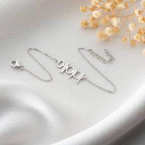 Pu&ograve; includere: Un delicato braccialetto d'argento con caratteri coreani. Il braccialetto ha una catena sottile e una chiusura. I caratteri coreani sono collegati alla catena. Il braccialetto &egrave; esposto su una superficie bianca.