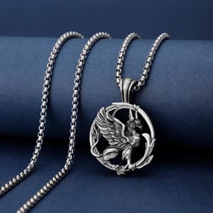 Könnte beinhalten: Silberfarbene Halskette mit einem Anhänger, der einen detaillierten Greifen in einem kreisförmigen Rahmen zeigt. Der Greif hat ausgebreitete Flügel und einen Löwenkörper. Die Halskette hat eine Box-Link-Kette. Der Hintergrund ist ein dunkles Blau.