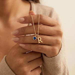 May include: A gold-colored necklace with a pendant featuring a blue heart-shaped stone and the letter "R". The necklace is held up by a person. The chain is delicate and the pendant is small.