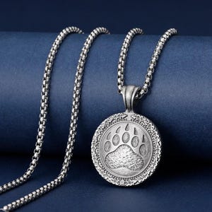 Könnte beinhalten: Silberfarbene Halskette mit einem runden Anhänger mit einem Bärenpfotenabdruck. Der Anhänger ist aufwendig detailliert und hängt an einer Kette. Die Halskette wird vor einem dunkelblauen Hintergrund präsentiert.