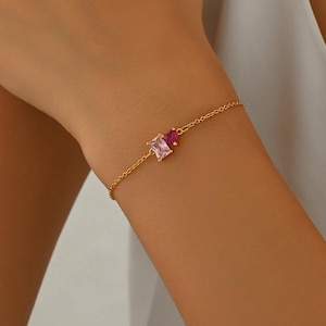 Puede incluir: Una delicada pulsera dorada con dos piedras preciosas de corte cuadrado en tonos rosas. La cadena de la pulsera es fina y elegante, con las piedras preciosas muy juntas. La pulsera se lleva en la muñeca.