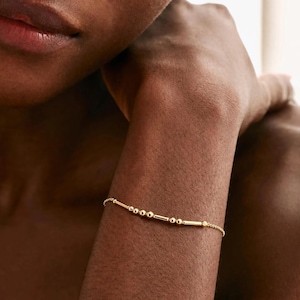 May include: A delicate gold chain bracelet featuring a series of small round beads and thin cylindrical bars arranged in a linear pattern. The metallic gold jewelry piece rests against the skin, showcasing a minimalist design with polished surfaces.
