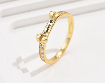 Puede incluir: Anillo de tono dorado con un diseño en forma de hueso. El hueso tiene una huella de pata y el nombre "Rocky" grabado. El anillo está adornado con piedras de circonita cúbica transparentes.