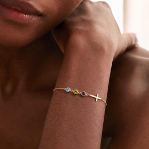 Puede incluir: Una delicada pulsera de cadena dorada con tres pequeñas piedras preciosas redondas engastadas en azul claro, verde lima y azul oscuro, seguida de un pequeño dije de cruz dorada lisa unido a la cadena, creando un accesorio de muñeca sencillo.