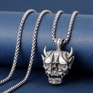 May include: A silver-colored pendant necklace featuring a detailed, horned demon skull design. The skull has sharp teeth and a grimacing expression. The chain is a linked, box-style design. The background is a solid blue.