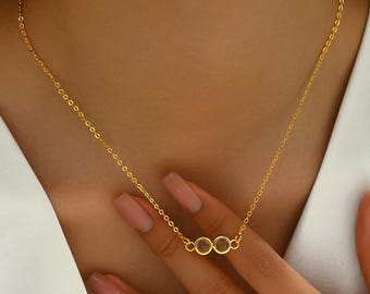 May include: A delicate gold necklace featuring three small, round, clear gemstones. The necklace has a thin chain and is displayed against a neutral background. The gemstones are set in gold bezels, creating a minimalist and elegant design.