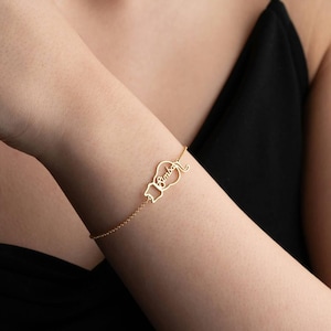 May include: A delicate gold bracelet featuring a cat silhouette with the name "Simba" inscribed within. The bracelet is attached to a fine chain, creating a minimalist design. The bracelet is worn on a wrist.