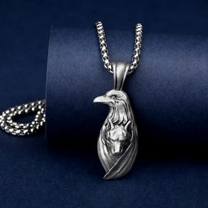 Può includere: Collana con pendente argentato raffigurante un'aquila con la testa di un lupo al centro. Il pendente è sospeso a una catena abbinata. Sfondo blu scuro.