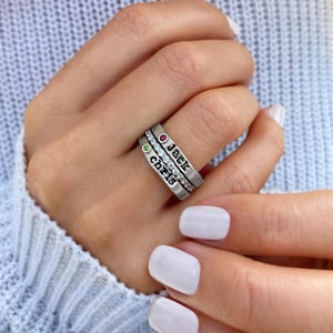 May include: Three silver rings stacked on a finger. The top ring has a red gemstone and the name "Jack". The middle ring is encrusted with small clear stones. The bottom ring has a green gemstone and the name "Chris".