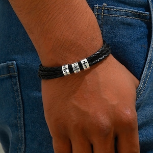 May include: Black leather bracelet with silver beads engraved with the names "Mae", "Logan", and "Autumn".