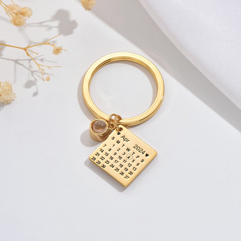 May include: Gold-toned keychain with a small square charm featuring a calendar for April 2024 with the number 4 highlighted. The charm also has a small clear crystal.